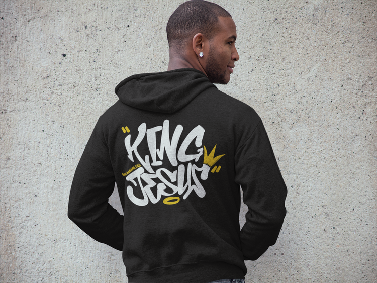 An african american man showing the back of a black hoodie with a grafitti style lettering of the words King Jesus, with a crown on the end of the letter S on a concrete background.