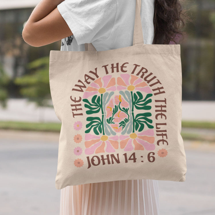 A woman in the streets carrying on her shoulder a natural color tote bag with the bible verse John 14:6, and the words saying "The way the truth and the life" with a very matisse style flower design in the center of the bag.