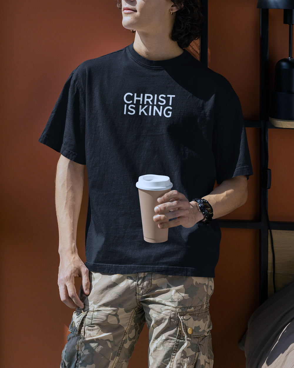 a young man holding a cup of coffee wearing an oversized box t shirt that is black with the words Christ is King