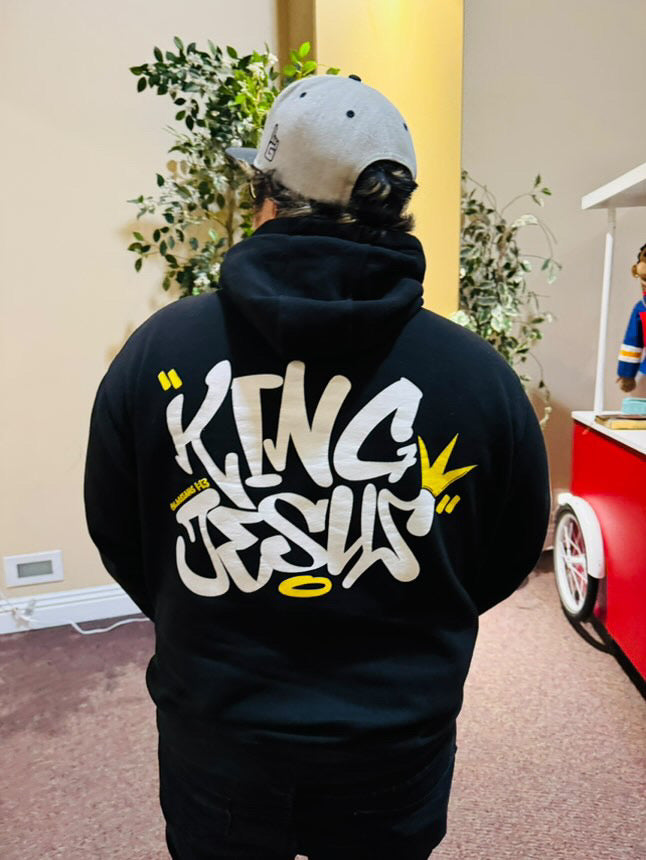 asian man wearing a King Jesus graffitis style hoodie shown on the back, wearing a baseball cap and black jeans.